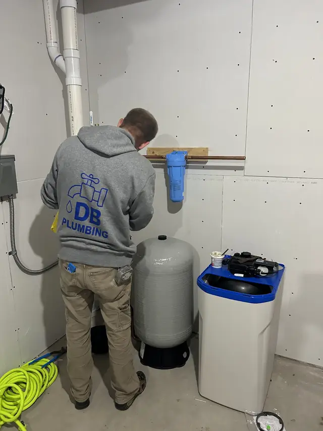 A DB Plumbing technician in a grey hoodie printed with the blue DB Plumbing faucet logo, working at a new pressure tank beside a whole-house filter and water softener in an unfinished basement