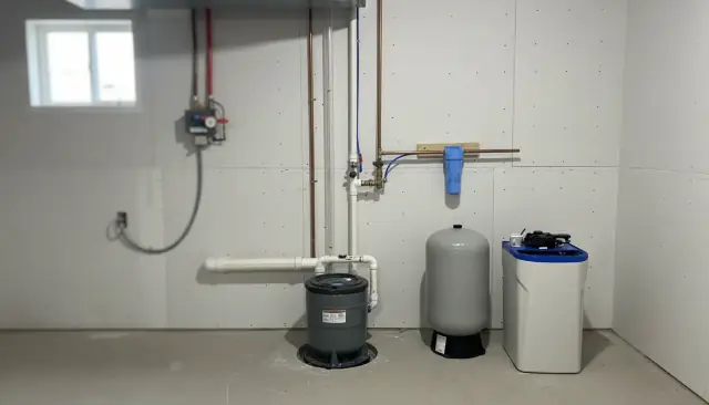 A basement corner with a covered sump basin beside a pressure tank and softener